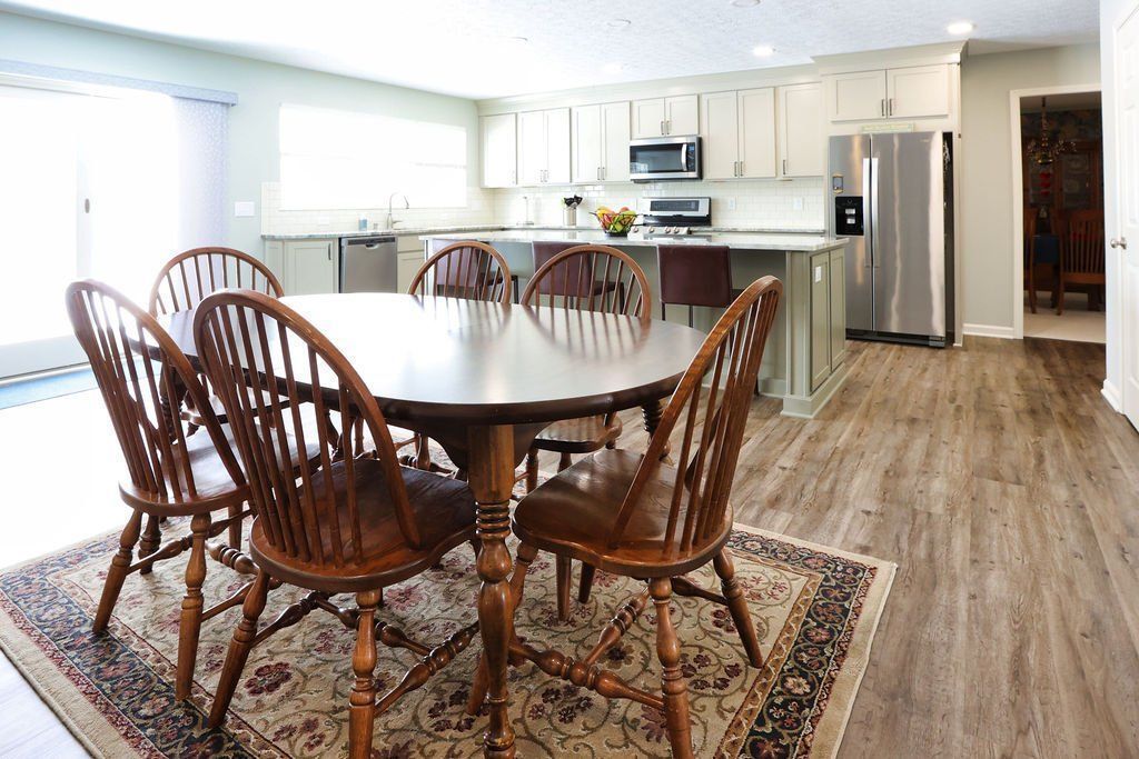Bright dining room with oval wooden table, chairs, patterned rug, and open kitchen with wood floors