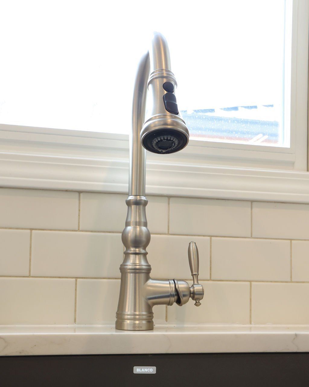 Silver kitchen faucet with pull-down sprayer, mounted on white tiled backsplash in front of a window.
