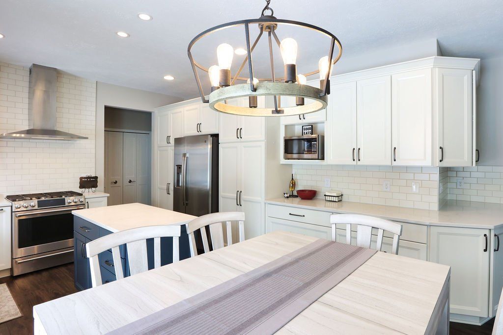 Spacious white kitchen with island, table, stainless steel appliances, and a decorative light fixture.