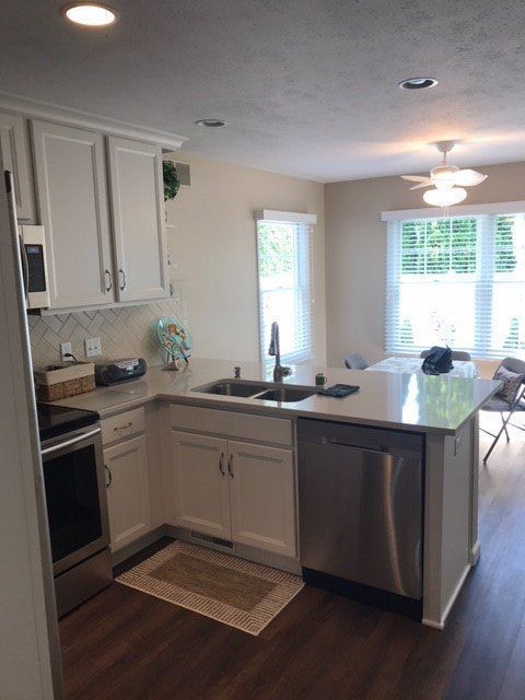 Bright kitchen with white cabinets, dark countertops, stainless dishwasher, and a sink by a window.