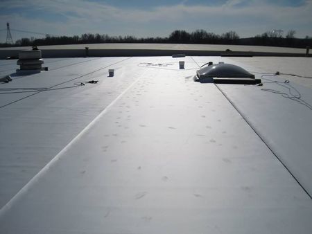 Flat, light-colored commercial roof with skylight, vents, and safety lines under a bright blue sky.