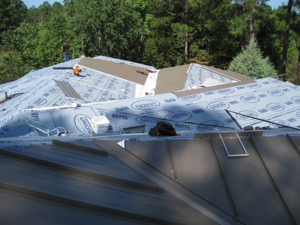Roofing work in progress; metal roof partially installed; blue underlayment visible; worker on roof in daylight.