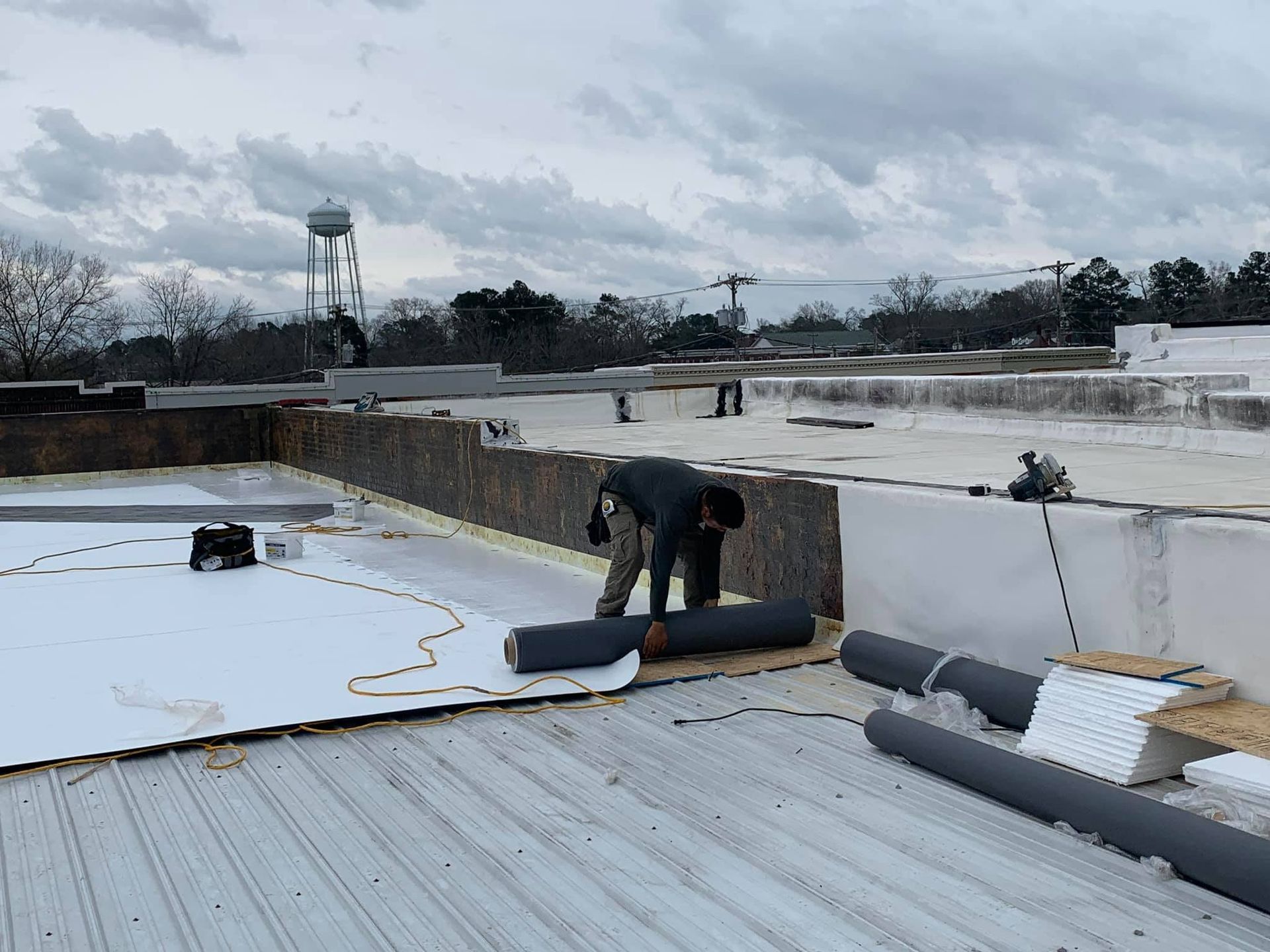 Roofer rolling out a dark membrane on a flat roof. A water tower is in the background on an overcast day.