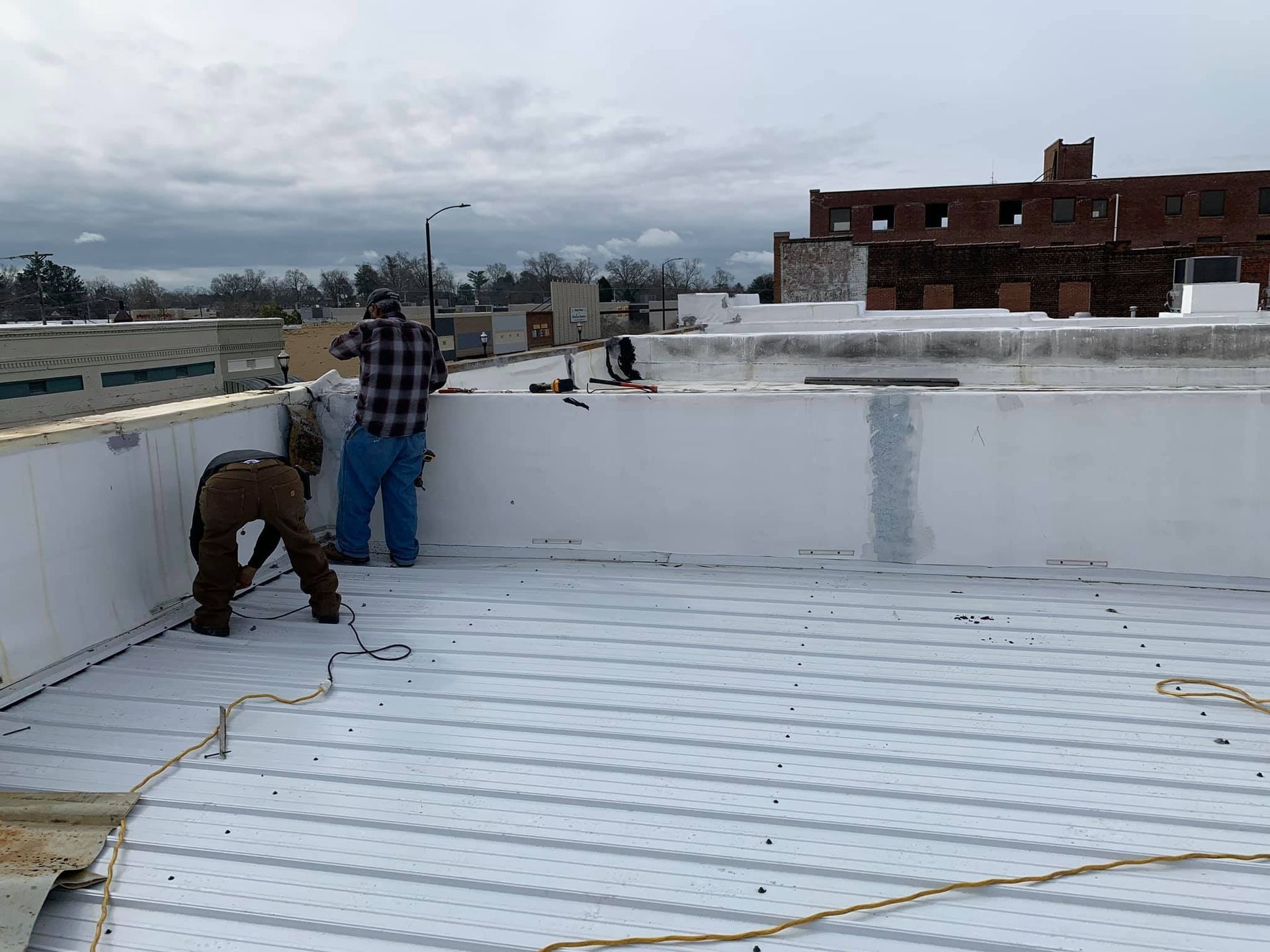 Two workers installing roofing materials on a flat roof under an overcast sky.
