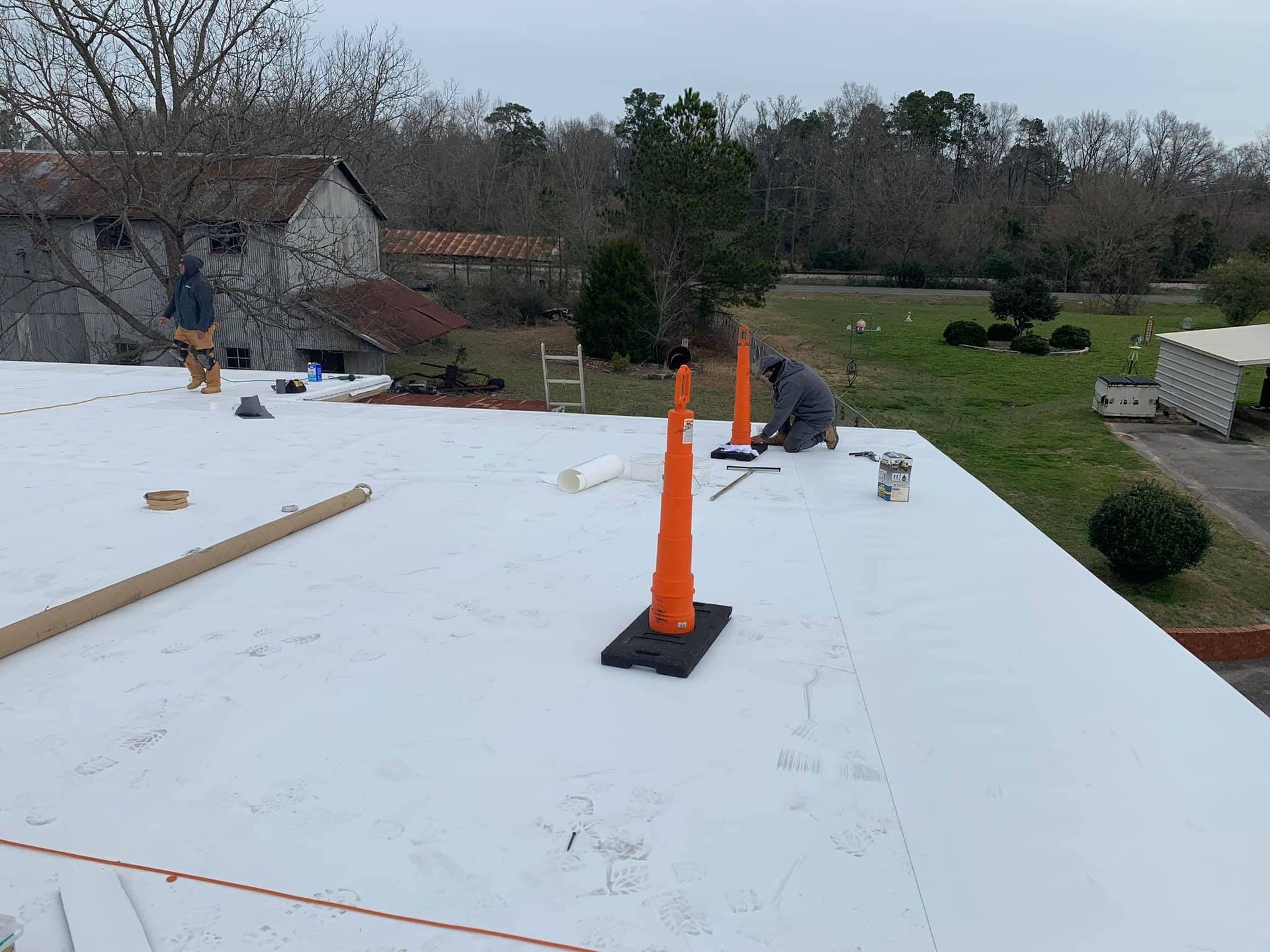 Workers on a flat roof, installing white roofing material. Orange cones, trees, and buildings visible in the background.