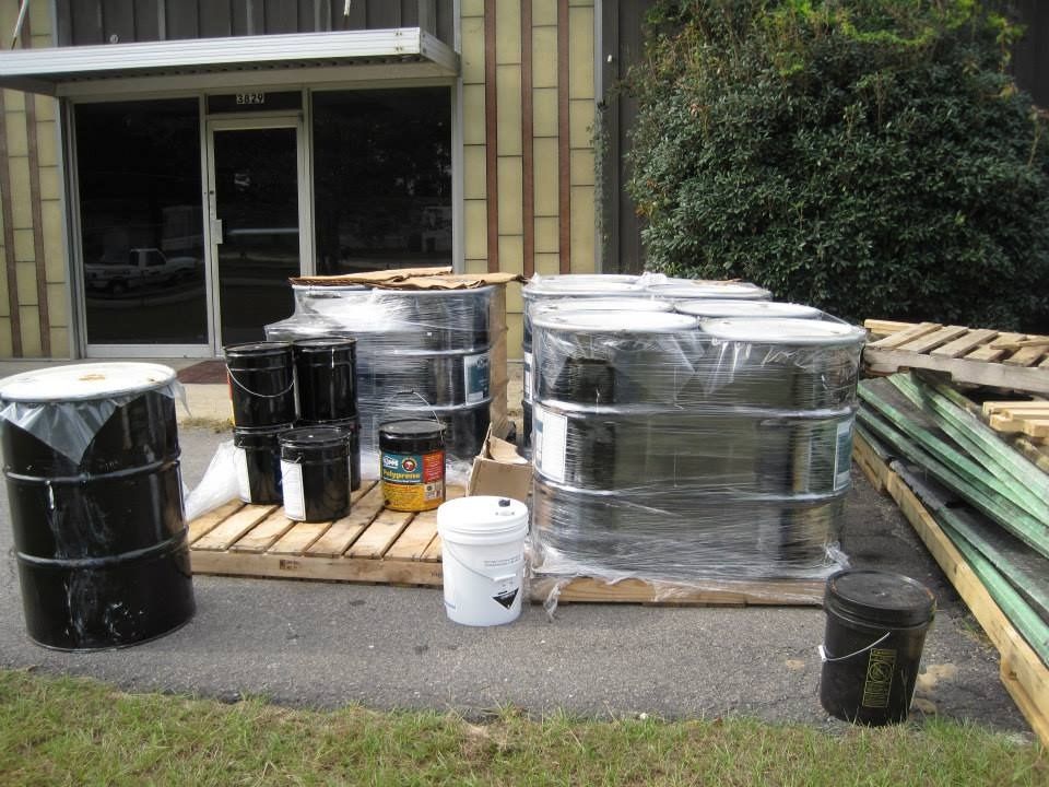 Black barrels and buckets on pallets outside a building with a glass door and brown exterior.