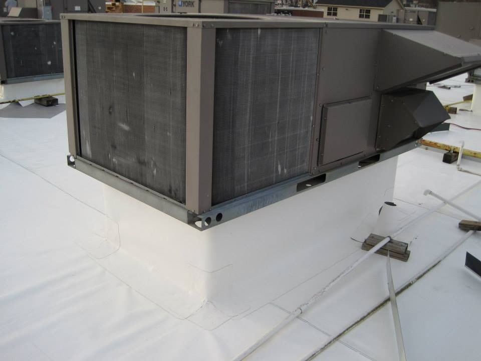 Rooftop HVAC unit on a white membrane roof. Gray metal unit with fins, vents, and ductwork.