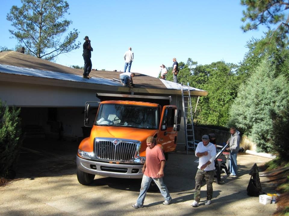 Roofing crew on a house; men on roof, orange truck in driveway, sunny day.