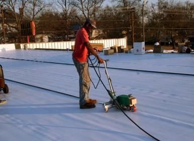 Man using a machine to seal a white roof. The man wears a red shirt and camouflage pants.