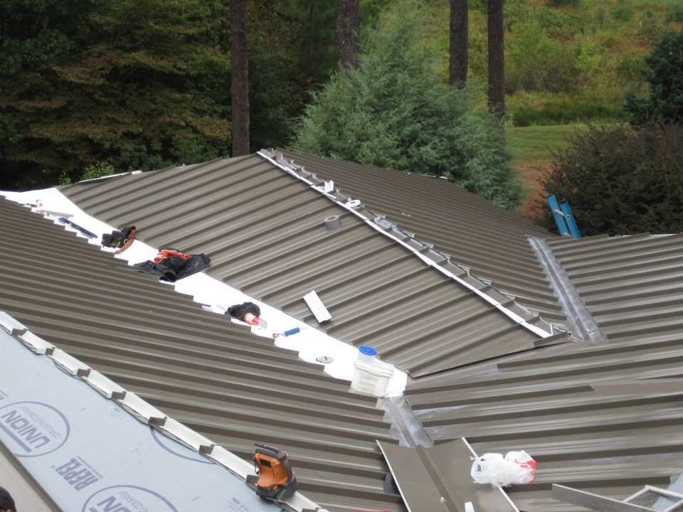 Metal roof being installed, tools, materials, and white underlayment visible. Green trees in the background.
