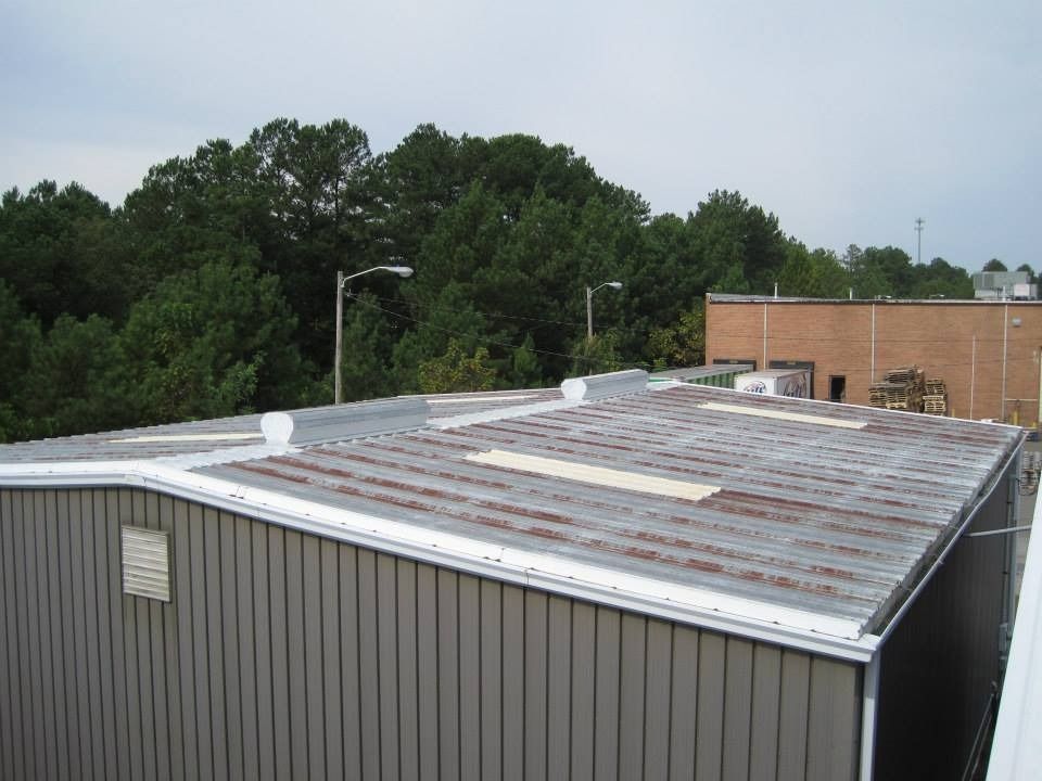 Gray metal building with a weathered, rust-colored roof, surrounded by trees and a cloudy sky.