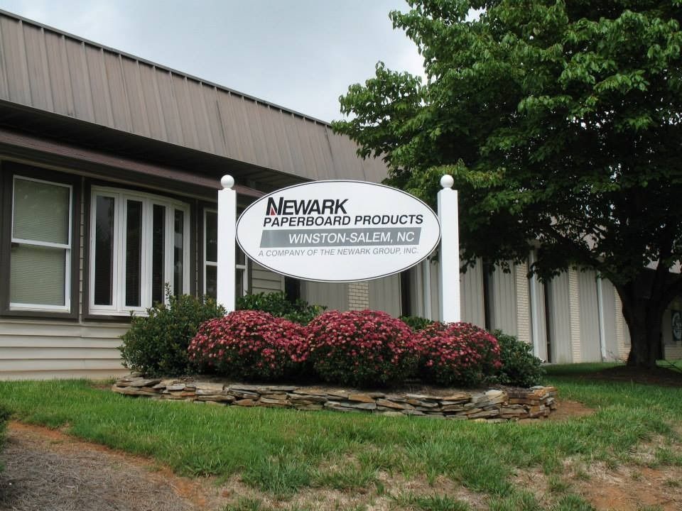 Sign for Newmark Wallboard Products in Winston-Salem, NC, with shrubs and a tree in front.