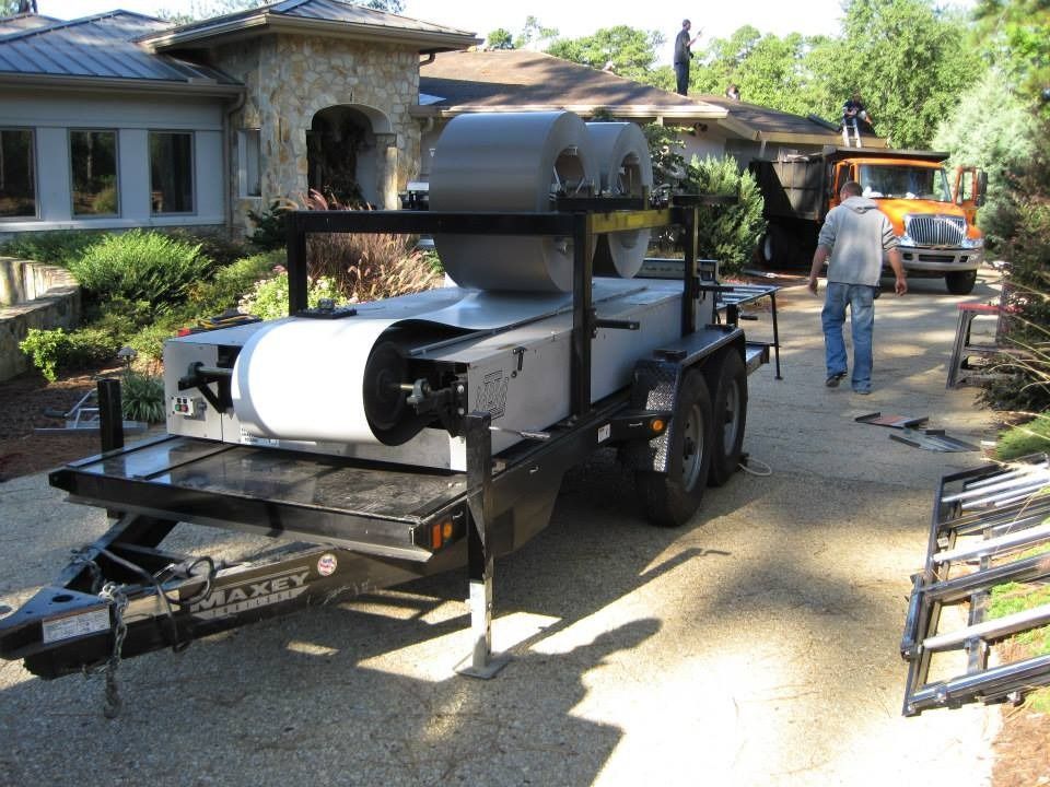 Trailer with metal roofing materials near a house, workers on the roof, and a truck.