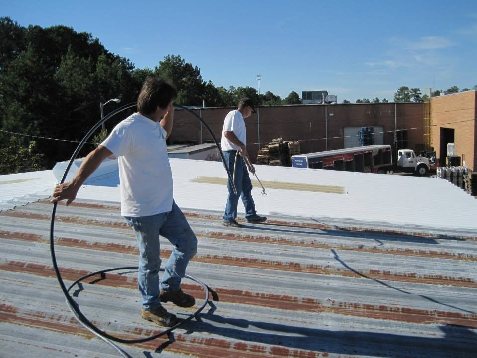 Two people on a rooftop, applying sealant. One holds a curved tool, the other uses a brush.