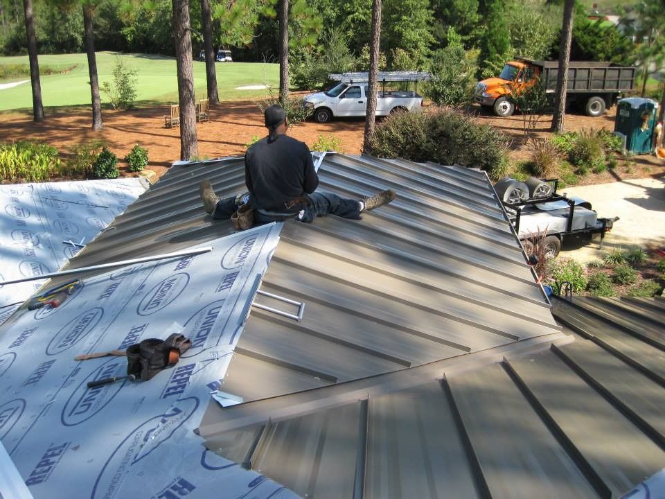 Person working on a metal roof, outdoors. Green trees and golf course in background.
