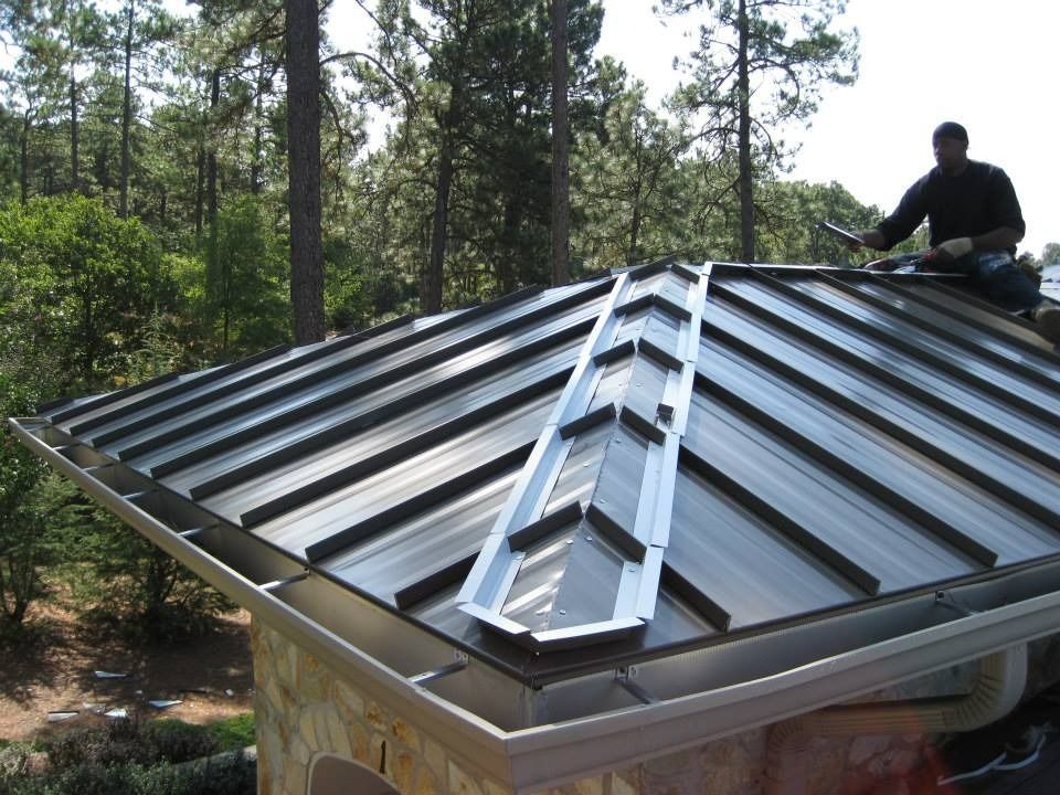 Man installing metal roofing on a house, sunny day, surrounded by trees.