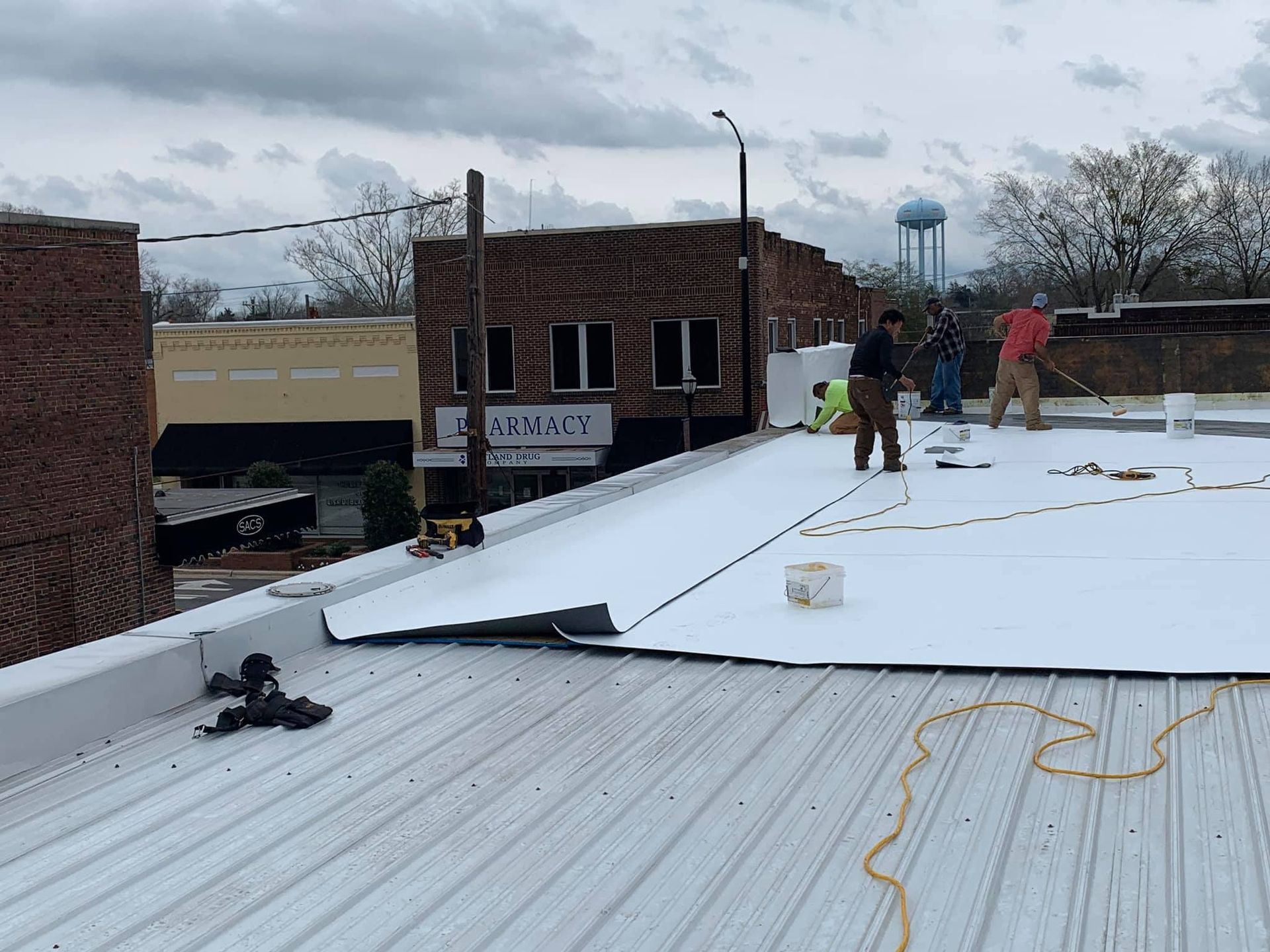 Workers applying material to a metal roof in a town with buildings and a water tower visible.