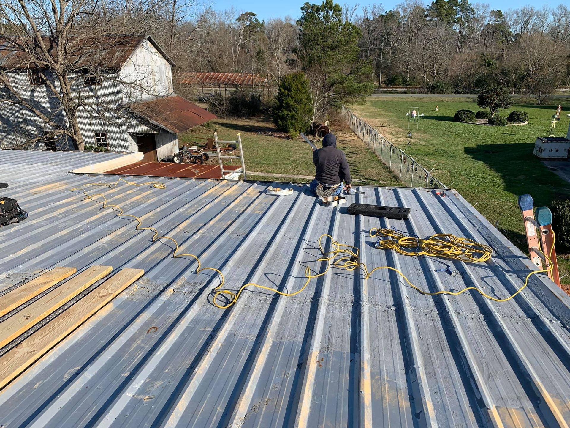 A person repairing a gray metal roof, with tools and materials laid out, and a rural background.