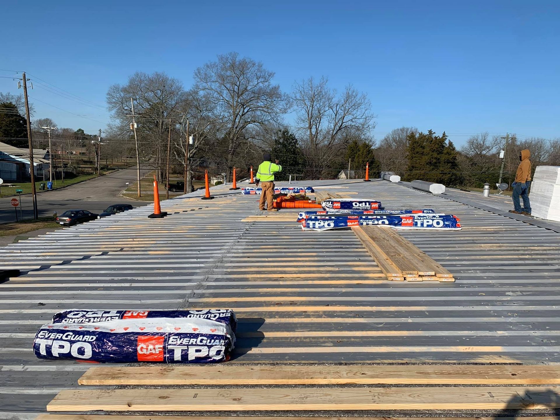 Workers installing roofing on a building; rolls of TPO membrane, wood, and cones are present.