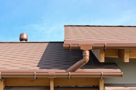 Brown roof with gutters and downspout, against a clear blue sky.