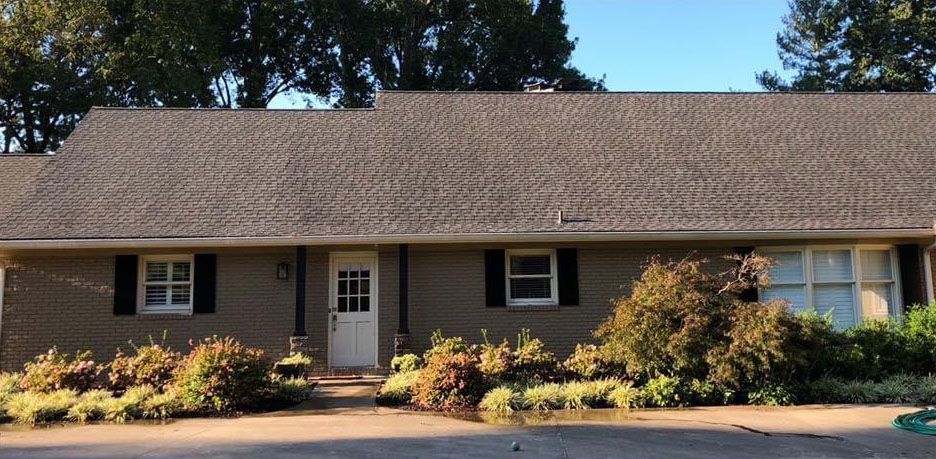 Low brick house with dark roof, white door, black shutters, and landscaping.