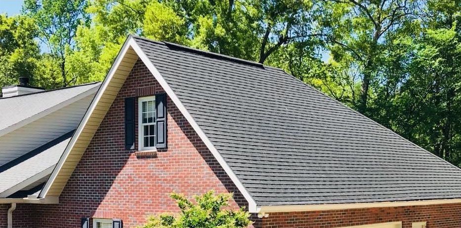 A brick house with a dark gray shingle roof against a backdrop of green trees.