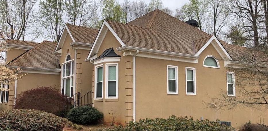 Beige house with brown roof, white trim, and windows. Trees in the background.