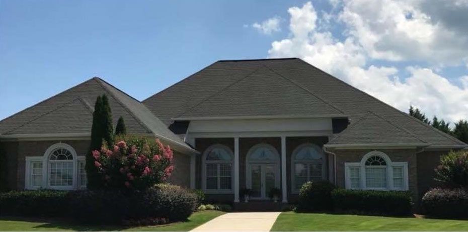 A large, beige house with a dark gray roof and a path leading to the entrance. Blue sky with some clouds.
