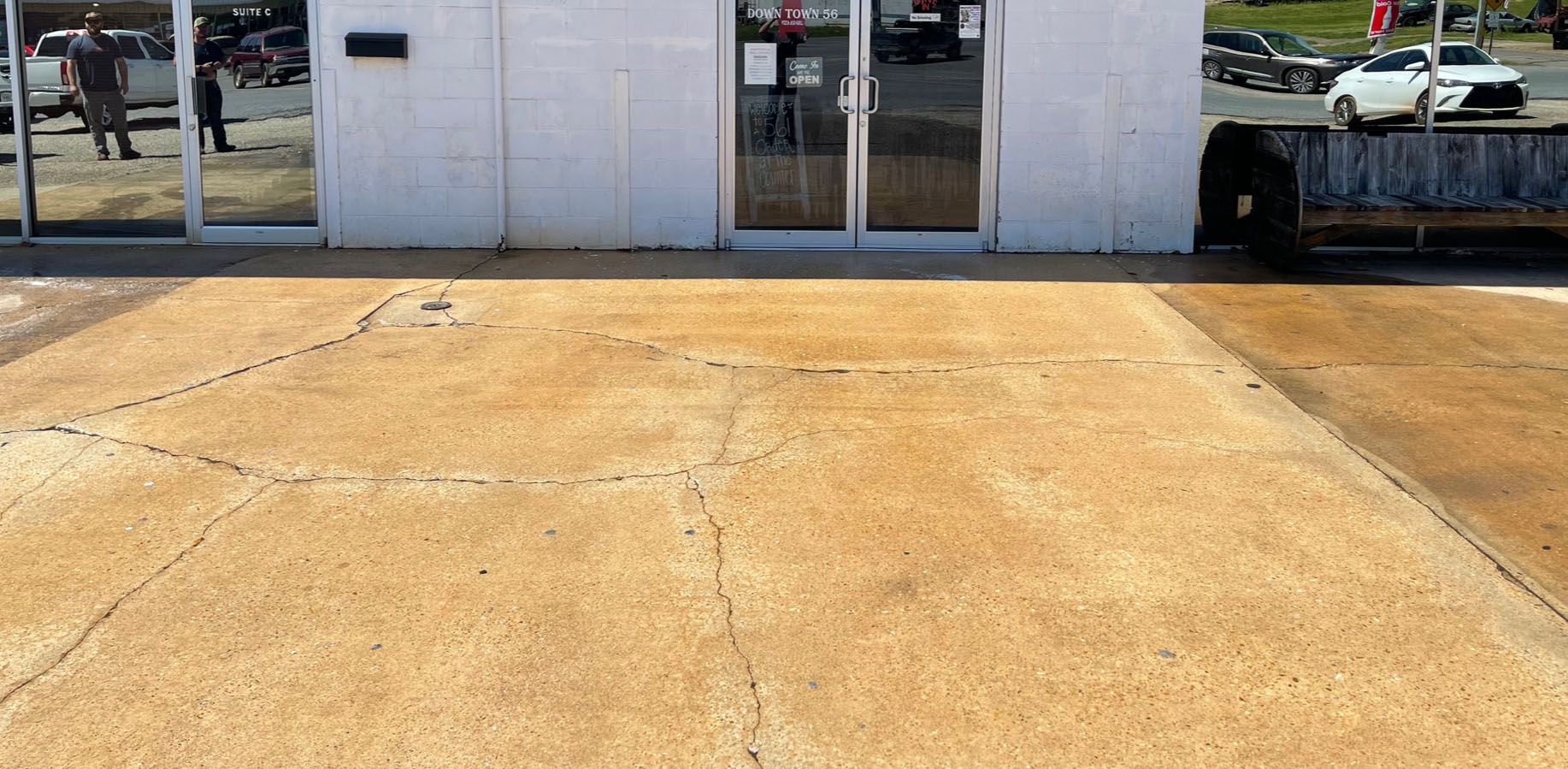 A concrete parking lot in front of a white building. Double glass doors are centered. Cars are parked nearby.