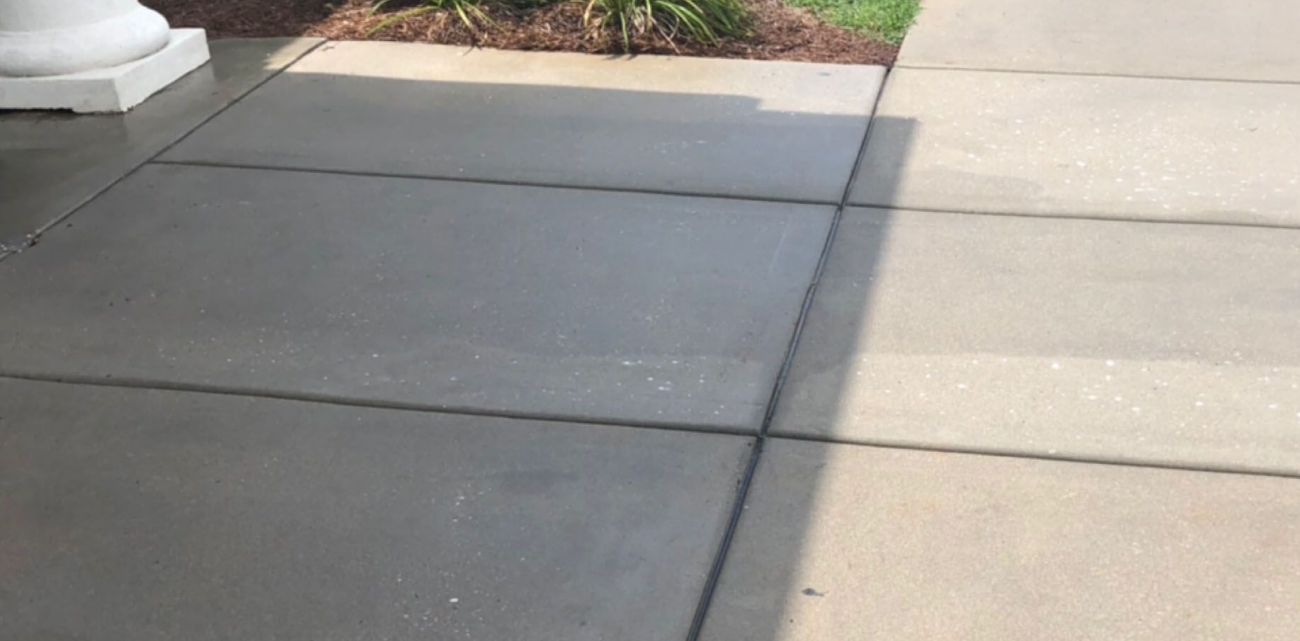 Sidewalk with concrete slabs. Sunlight casts shadows. Grass and a column are visible.