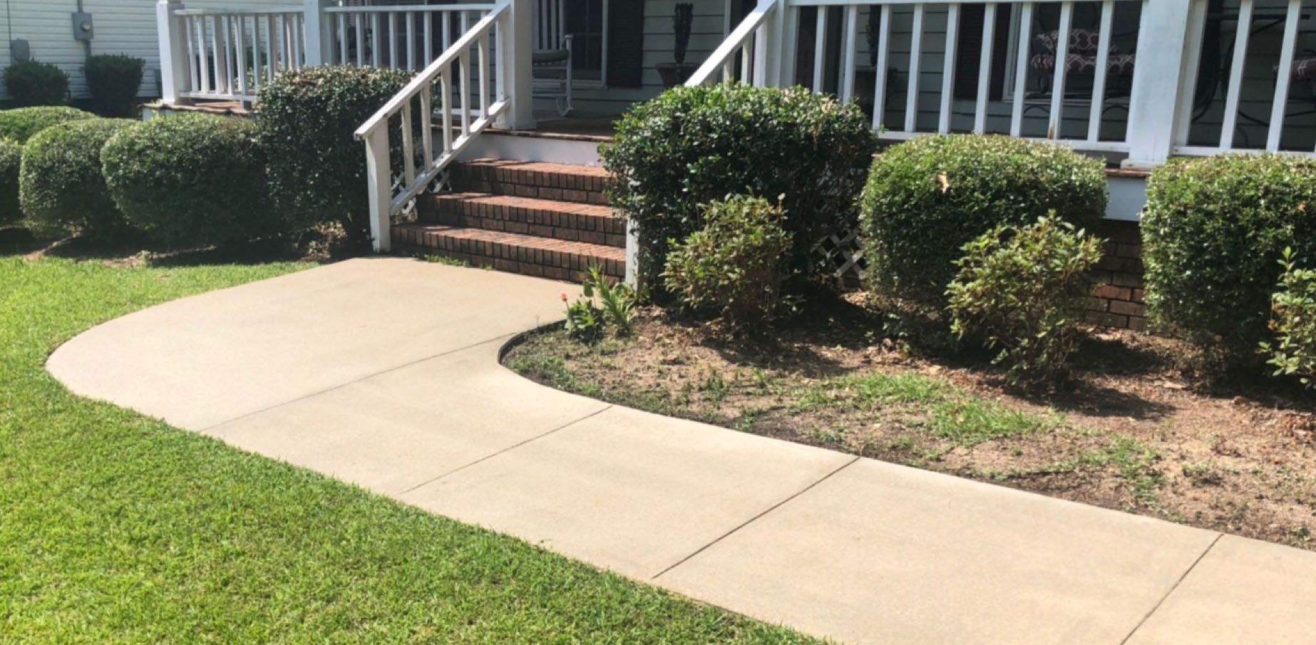 A concrete walkway curves towards a house with white railings, stairs, and green bushes.