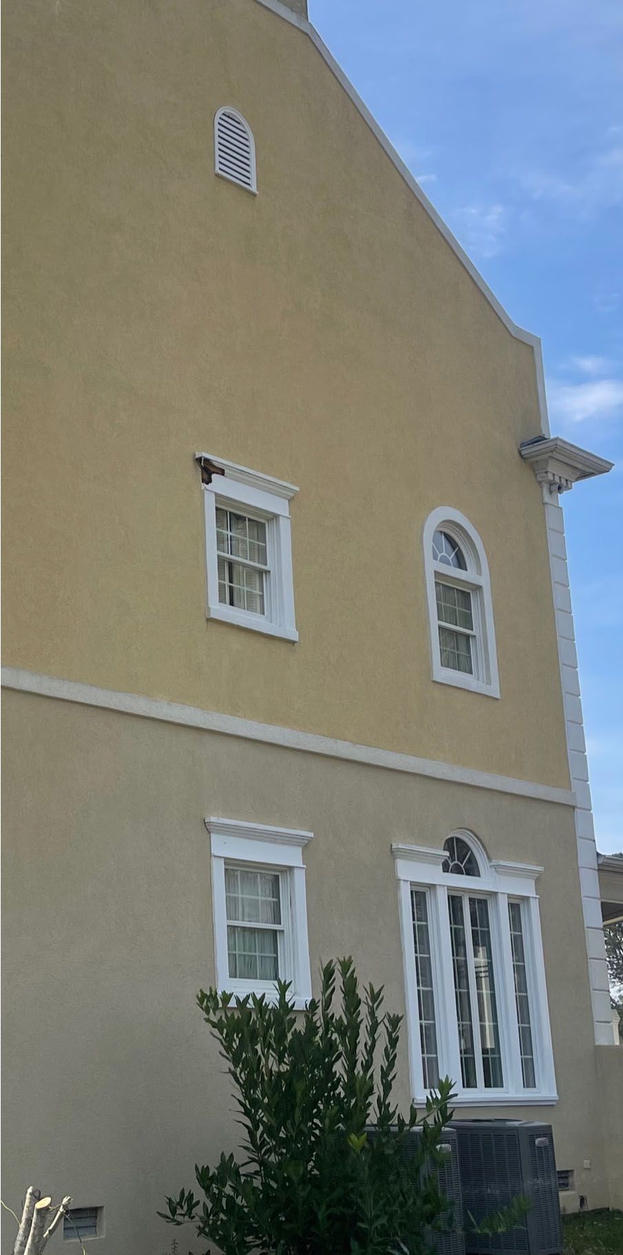 A two-story beige stucco building with white trim, windows, and a shrub in front, against a blue sky.