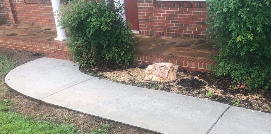 Concrete sidewalk curves past green bushes, a brick wall, and a large rock in front of a house.