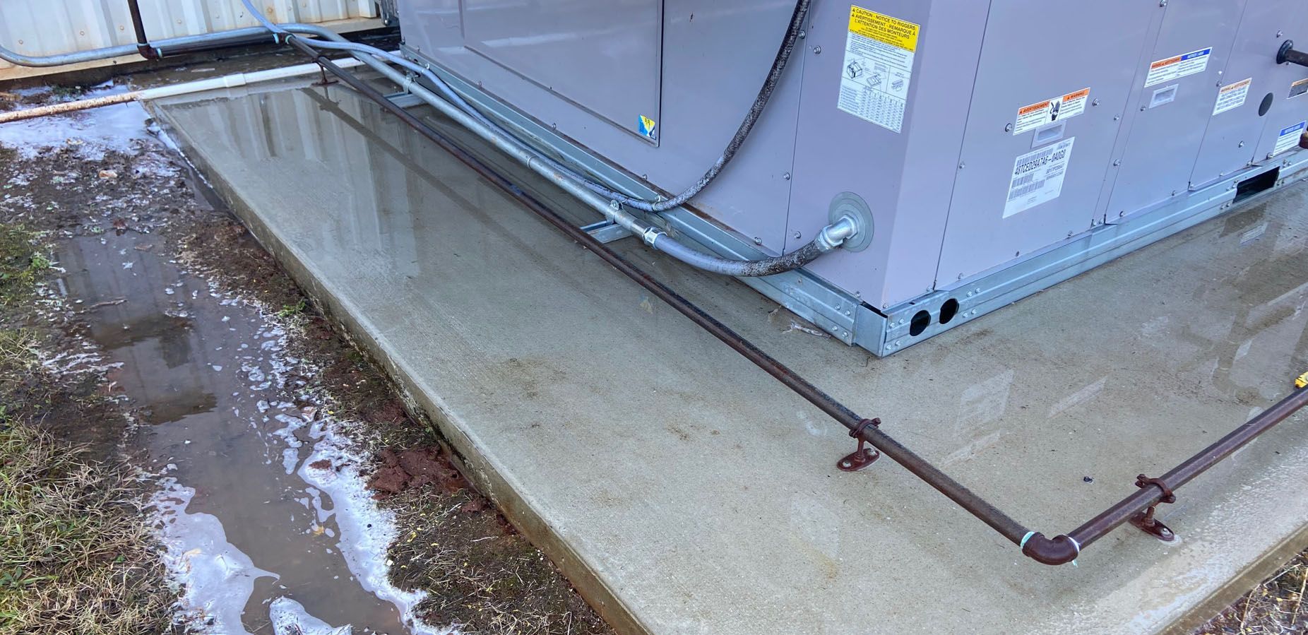 An AC unit on a concrete pad with brown pipes running along it. Ground is wet with patches of snow.