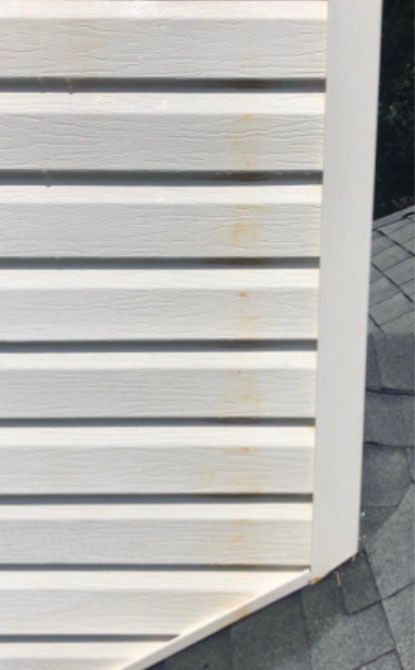 White vinyl siding with small brown stains, corner of a building with a dark gray roof.