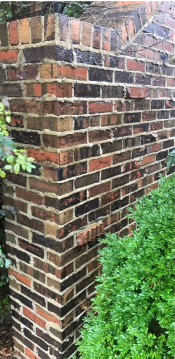 Brick corner of a building, various shades of brown bricks. Green bushes are in the foreground.