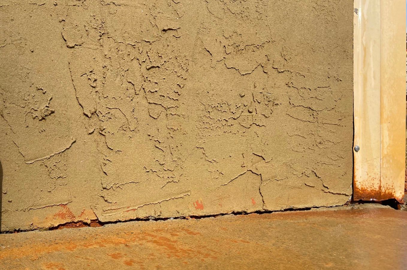 Textured tan stucco wall with cracks near the base, adjacent to a wooden support beam.