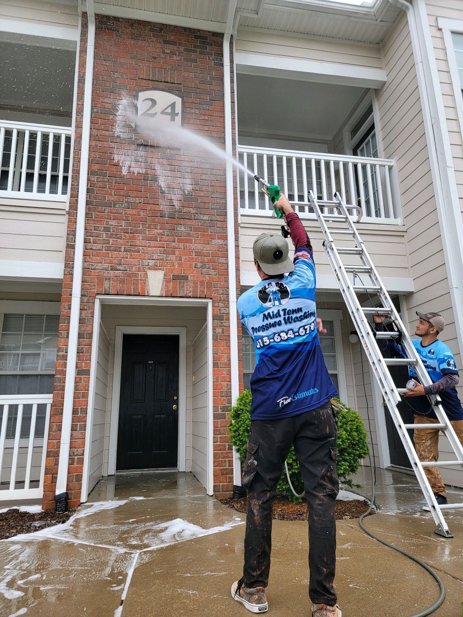 Two people pressure washing a building's brick facade. One on a ladder, other spraying near door