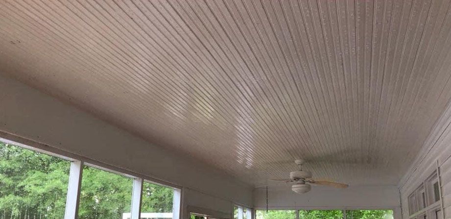 A porch ceiling with horizontal white wood paneling and a ceiling fan, with greenery visible through the windows.