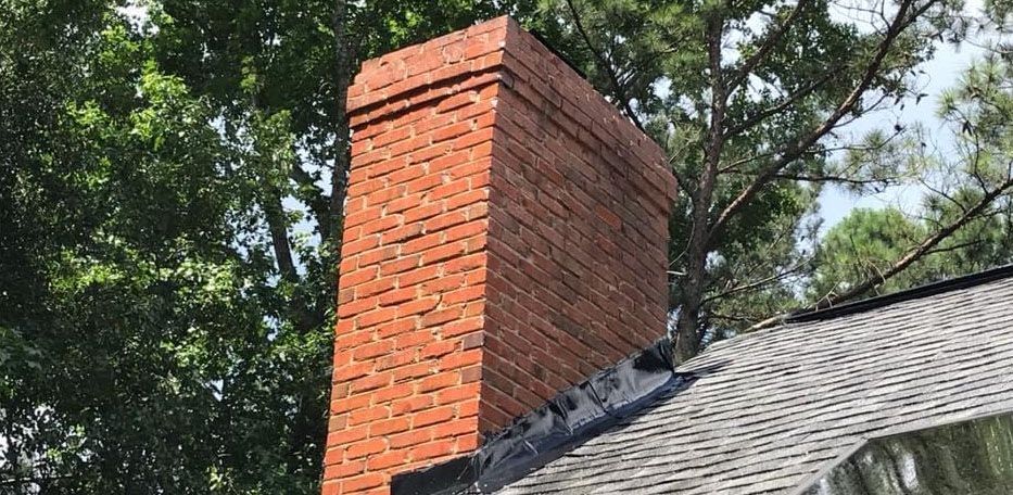 Brick chimney on a shingled roof, surrounded by trees.
