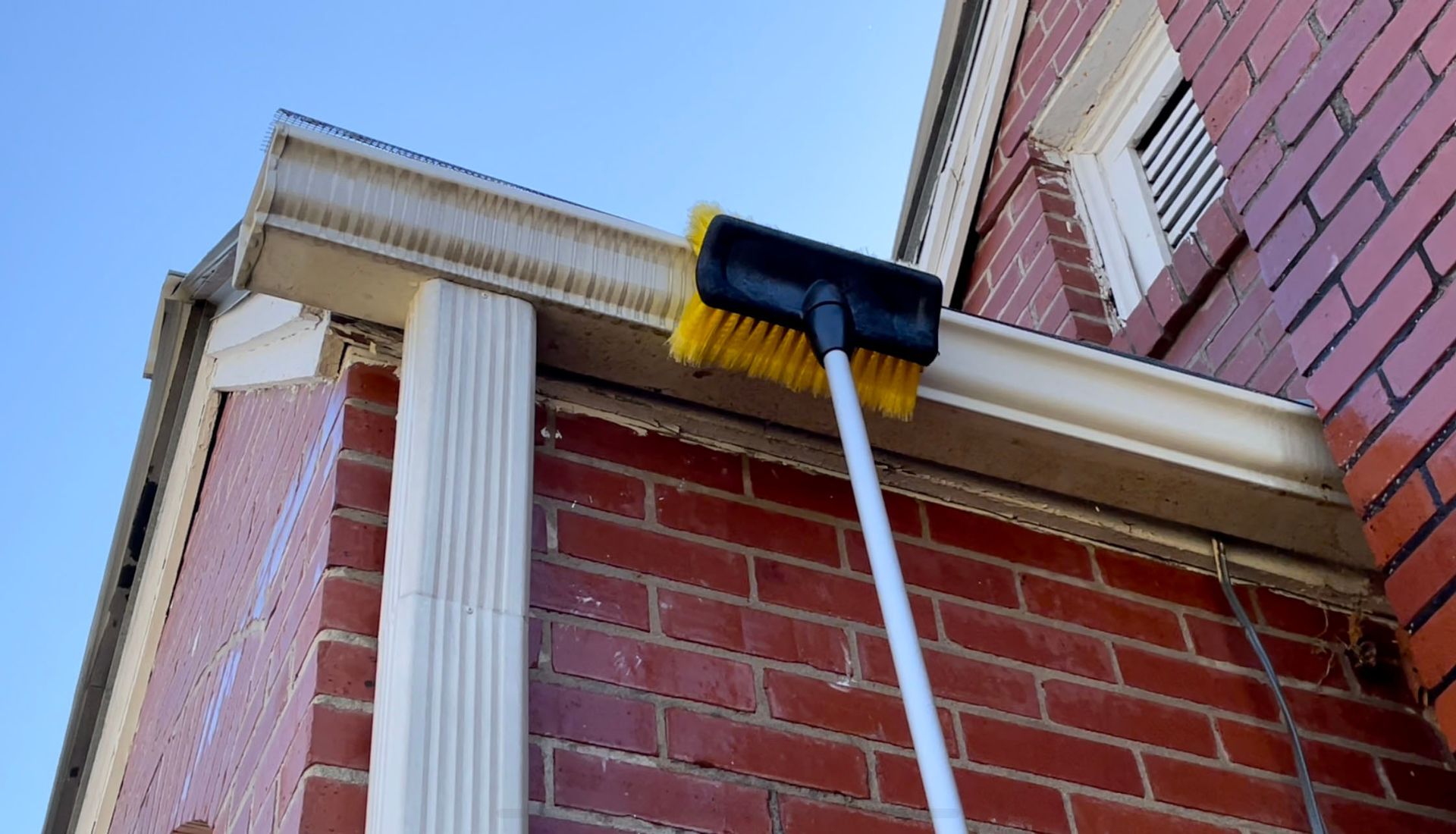 Cleaning a gutter with a long-handled brush on a red brick building against a blue sky.