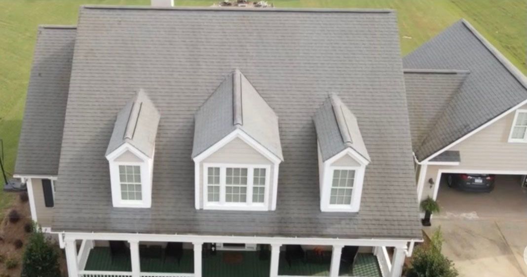 Aerial view of a house with gray roof, three dormers, and a porch. Green lawn in the background.