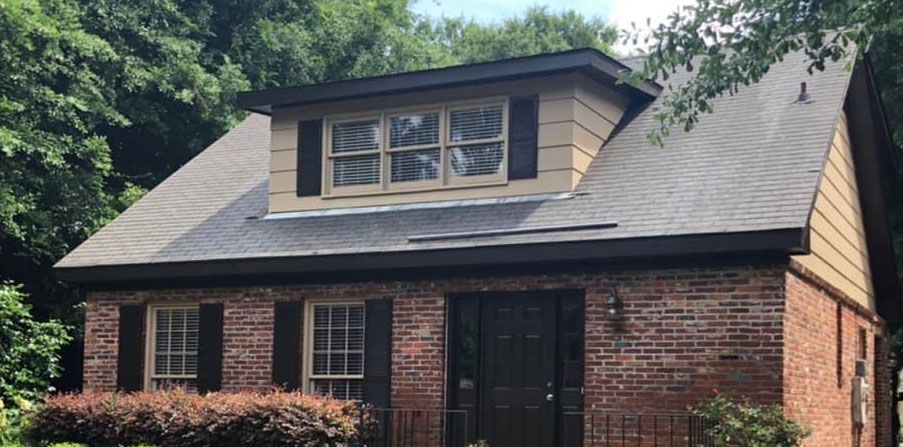 Brick house with dormer window, black shutters, and dark roof against green trees.