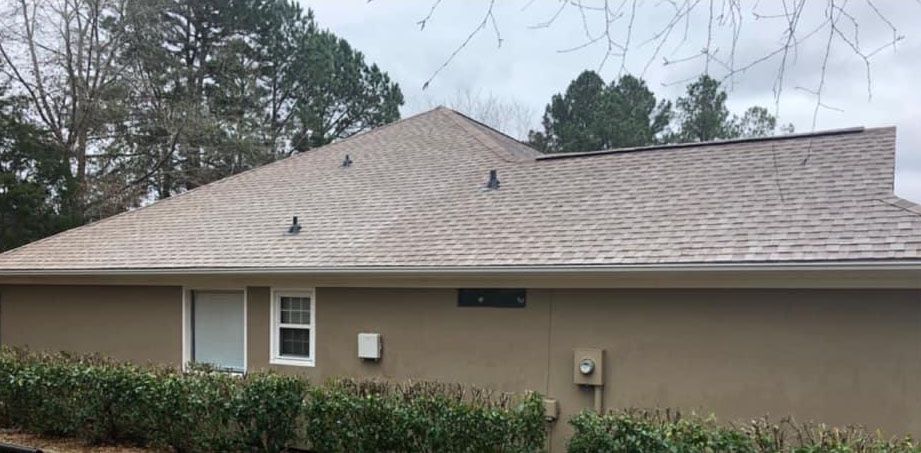 A beige-colored house with a brown roof, green bushes, and trees in the background.