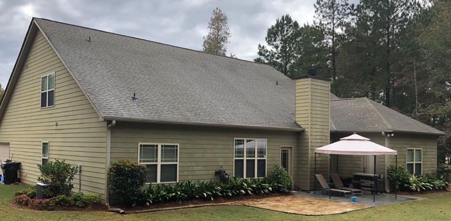 Back of a beige house with a dark roof, chimney, patio, and trees. Cloudy sky.