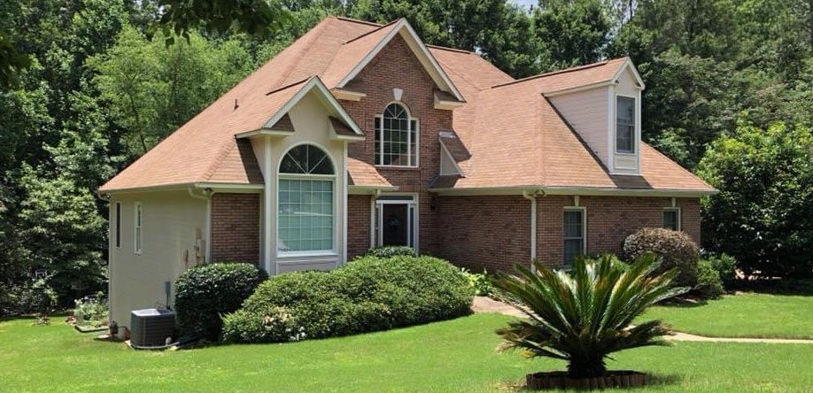 A two-story brick house with a brown roof, surrounded by green lawn, trees, and bushes.