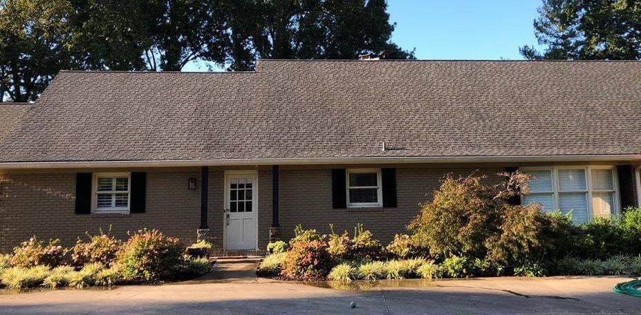 Exterior view of a one-story brick house with a brown shingled roof, white door, and landscaping.