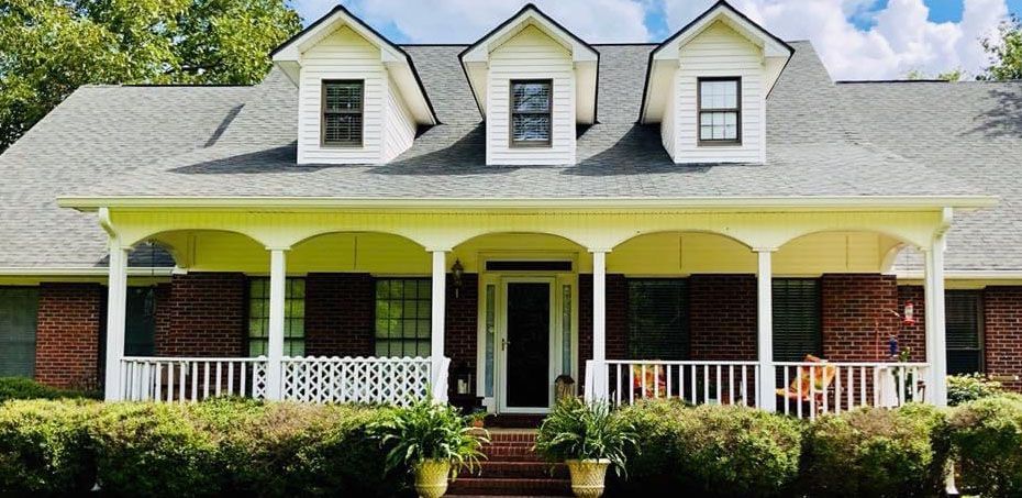 A brick house with a white porch, black shutters, and three dormers. Green foliage and a blue sky.