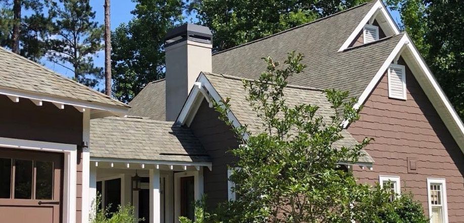 Brown house with gray shingle roof, chimney, and dormers, with trees in the background.