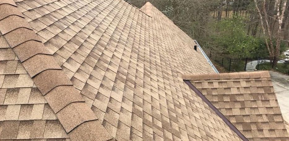 Tan asphalt shingle roof with visible patterns. A house exterior with tree background.